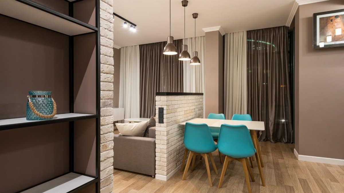 Chairs at table placed near white brick partition in living room with couch in modern apartment with decoration on shelf