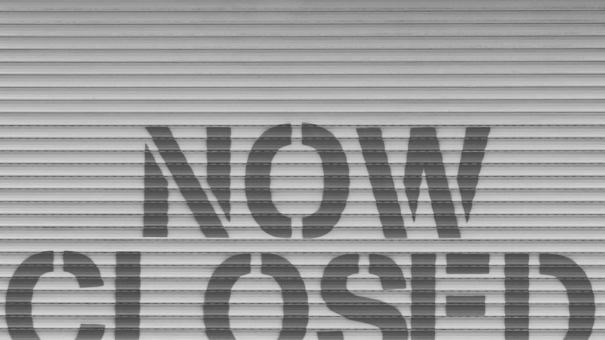 Black and white image of a 'Now Closed' sign on a steel shutter, symbolizing closure.