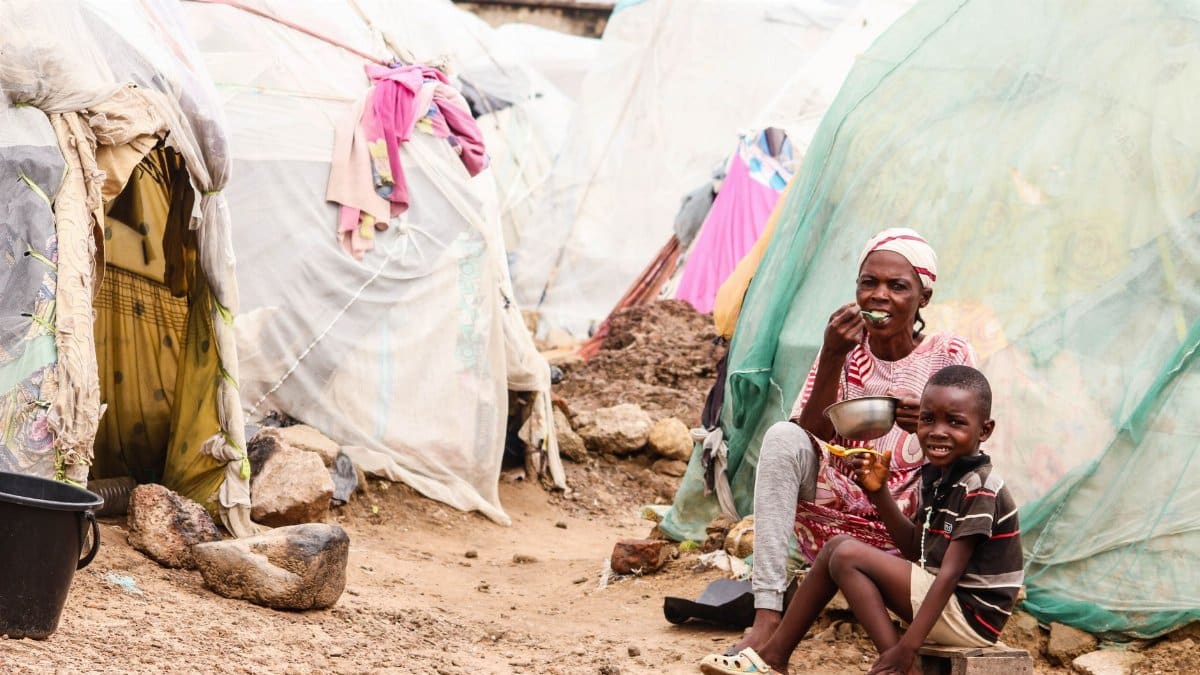 Mother and son enjoying a meal in a rustic village scene with makeshift housing.