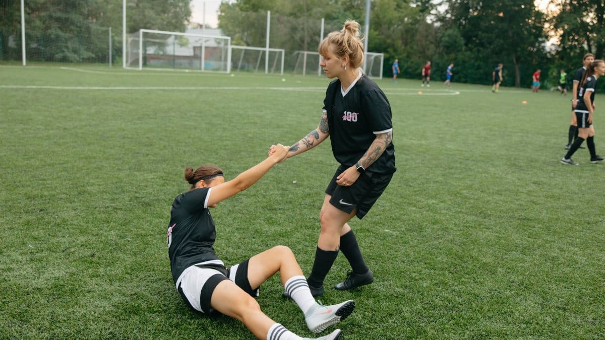 Two female soccer players in black uniforms on a field, one helping the other to stand up.