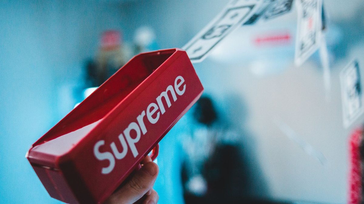 Close-up shot of a red toy gun labeled 'Supreme' shooting fake money, with a blurred background.