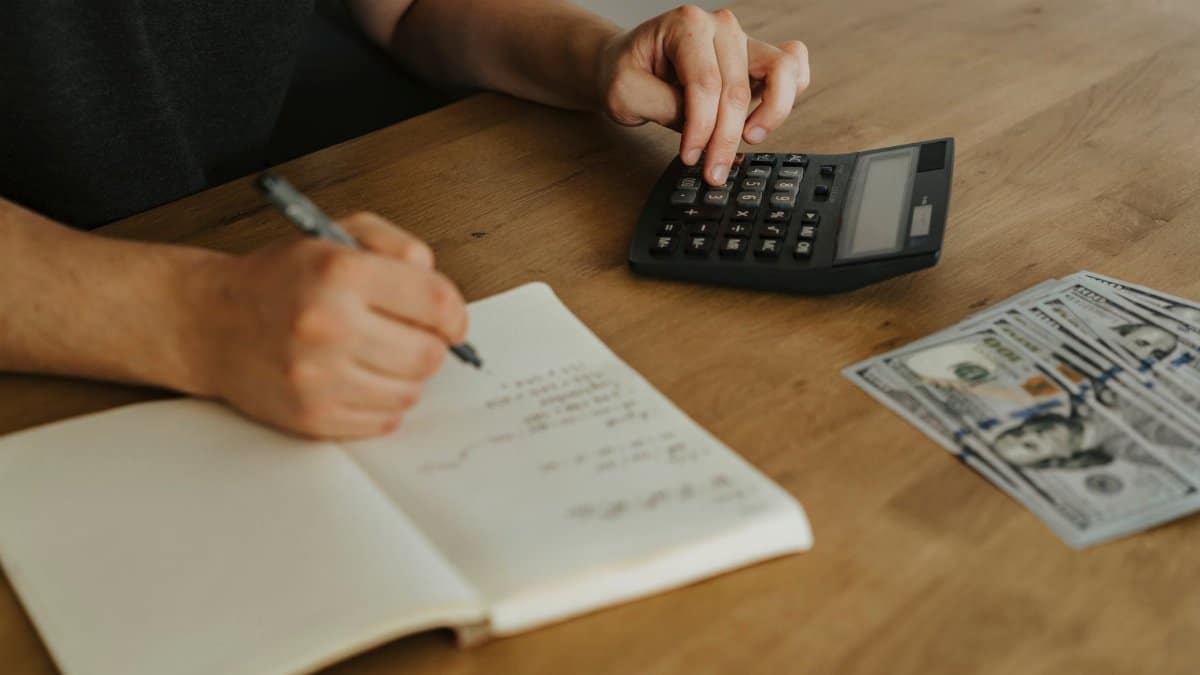 Close-up of hands using a calculator with notebook and cash on a wooden table.