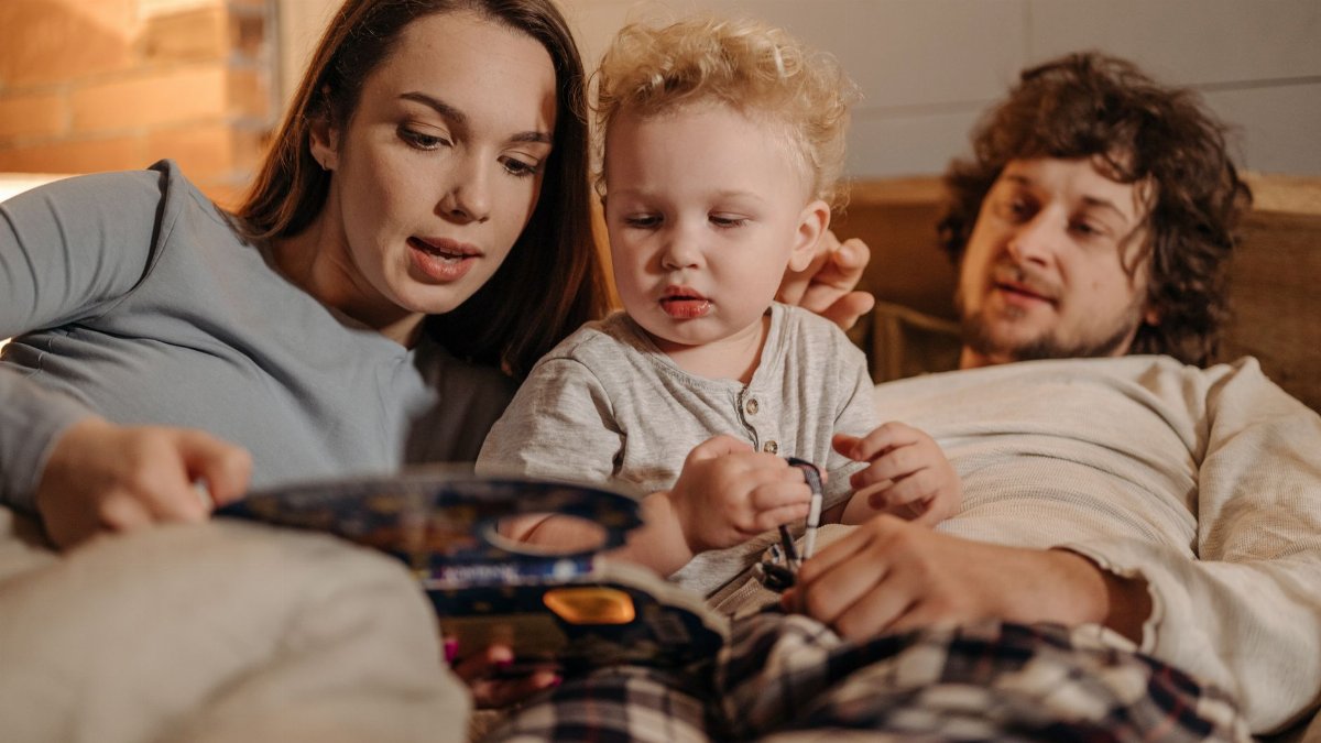 A family spending quality time in bed, reading a storybook to their toddler.