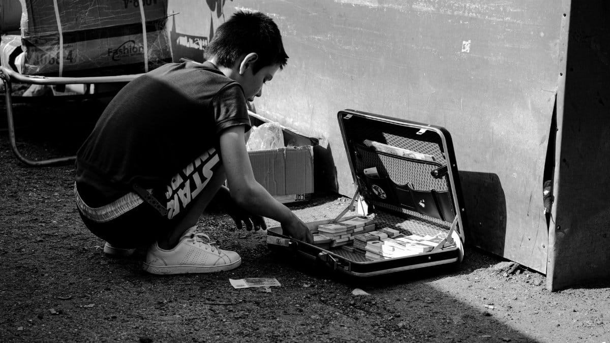 Black and white photo of a child examining money in a briefcase on a street.