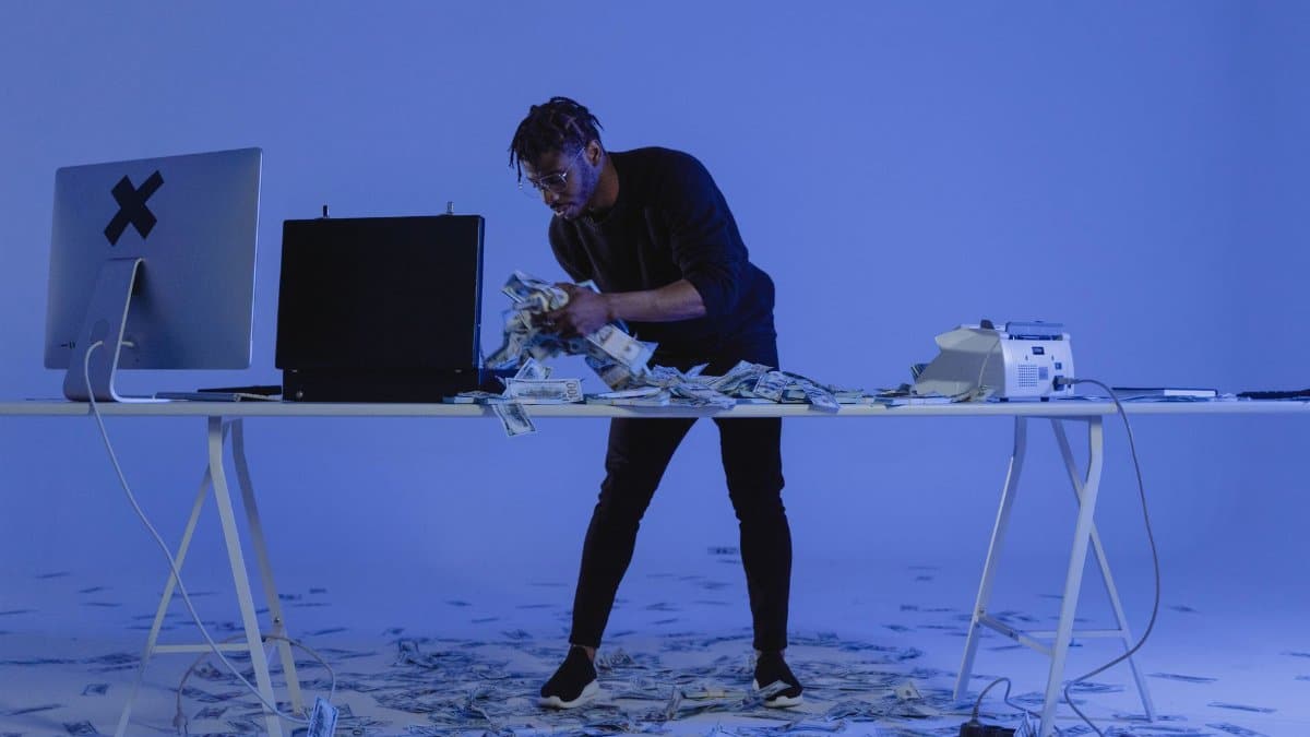 A man counting cash surrounded by computers and money in a blue-lit room, symbolizing cybercrime.