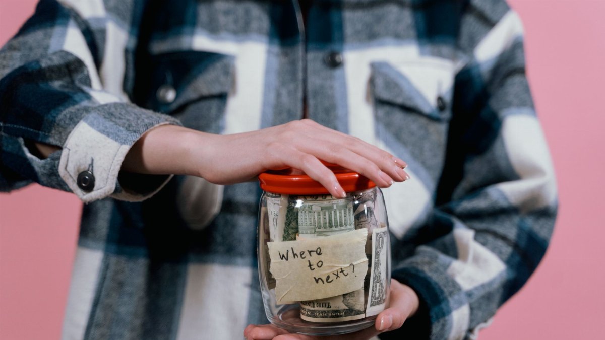 Close-up of a woman holding a savings jar labeled 'Where to next?' filled with US dollar bills.