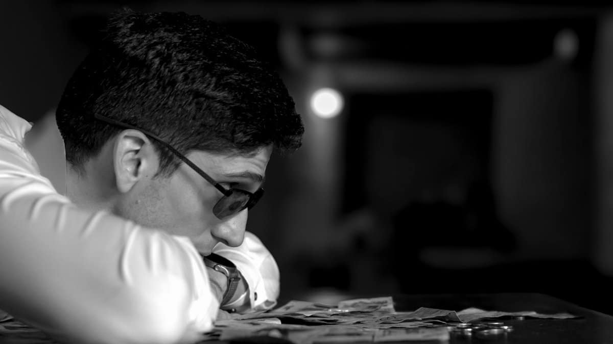 A man in glasses deep in thought, resting on a table with scattered money in a dimly lit room.