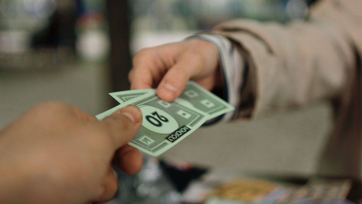 Close-up of hands exchanging Monopoly money outdoors in a park setting.