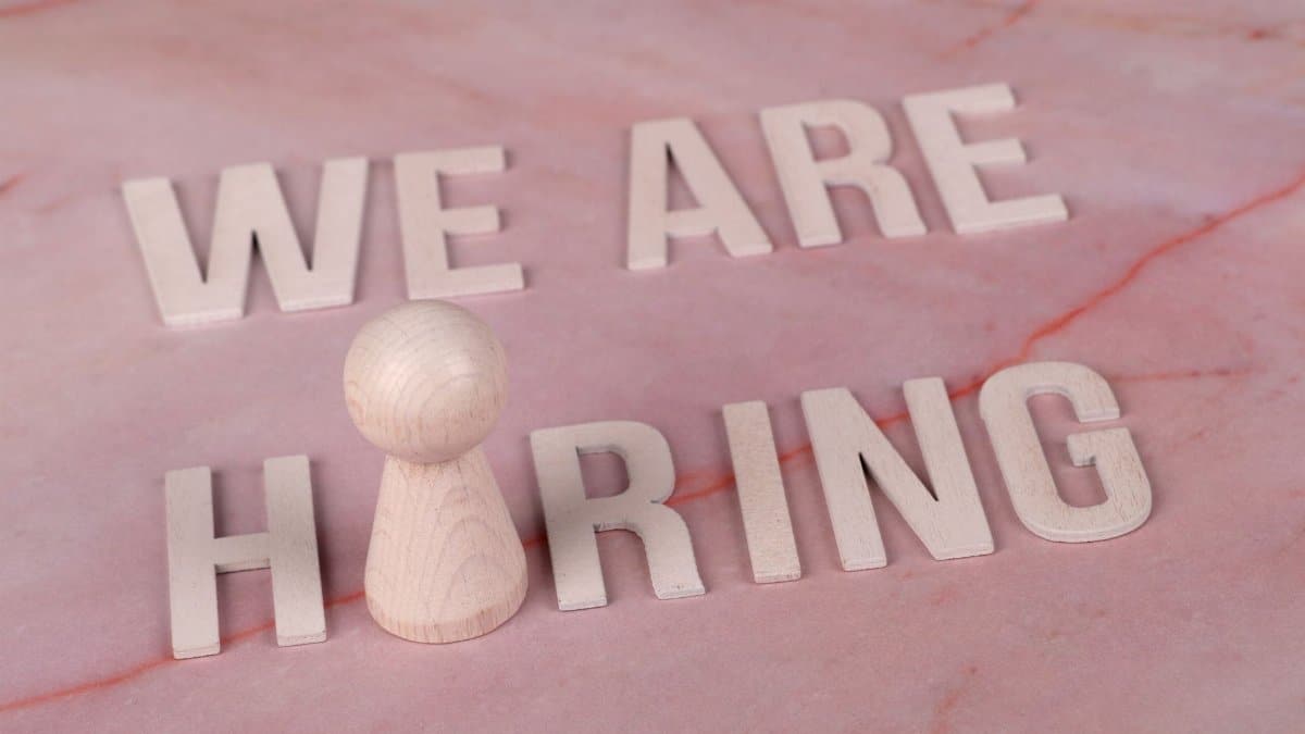 Wooden letters spelling 'We Are Hiring' on a pink marble surface with a pawn piece.