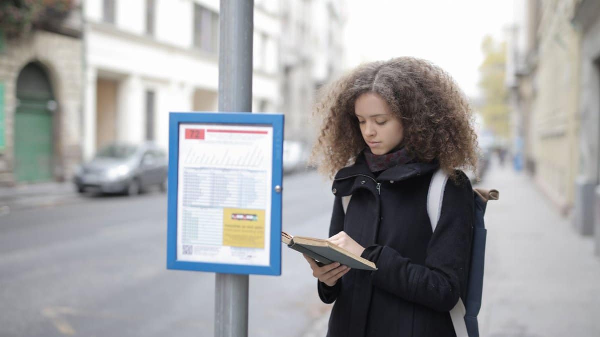A young woman with curly hair reads a book while standing at a bus stop on a city street.