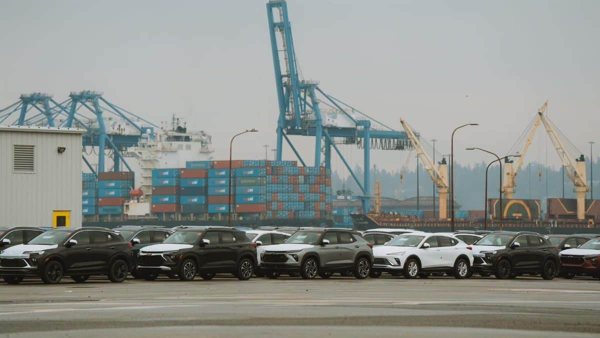 A lineup of parked cars at a bustling industrial shipping port with cranes and containers.
