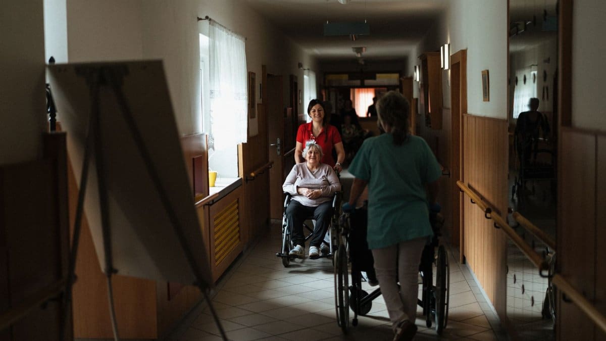 Caregiver assisting elderly woman in a wheelchair through a retirement home corridor.