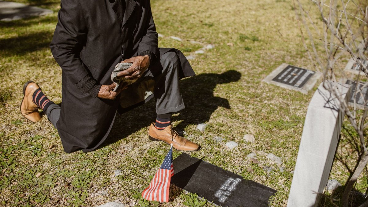 Man kneels by a grave with American flag, mourning peacefully outdoors.