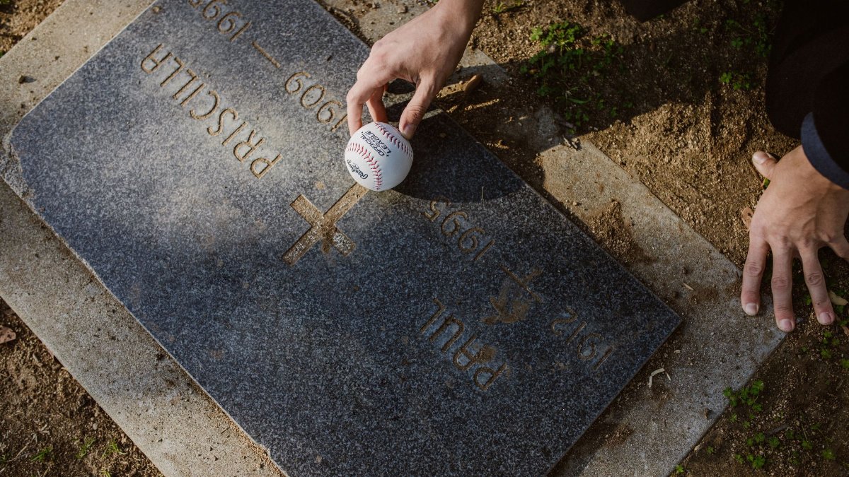 Hand placing a baseball on a grave's tombstone in a serene cemetery setting.