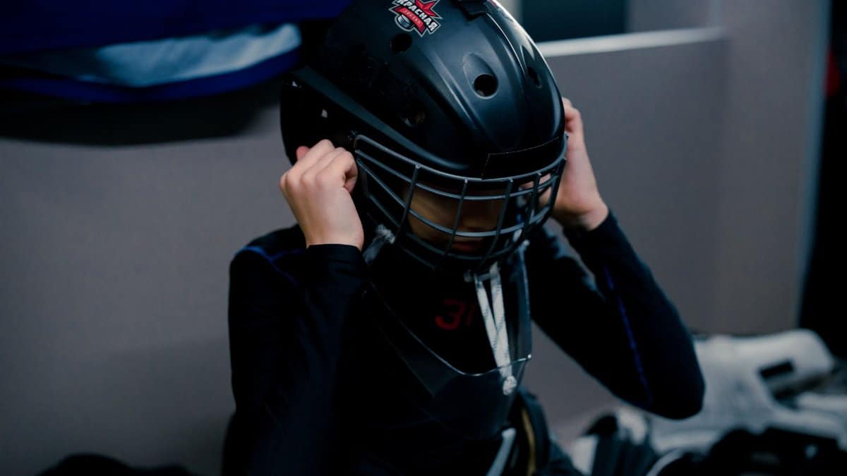 A teenage ice hockey goalie adjusts his helmet in a dressing room, preparing for a game.