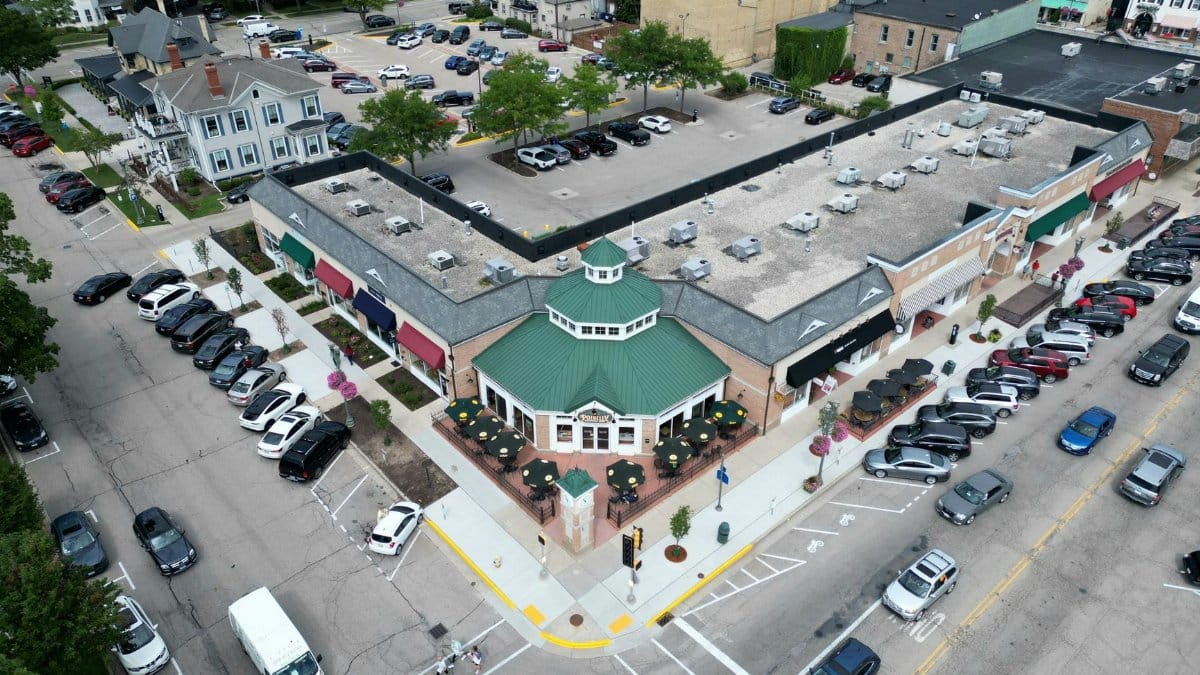 Bird's-eye view of a bustling suburban shopping center with parked cars and tree-lined streets.