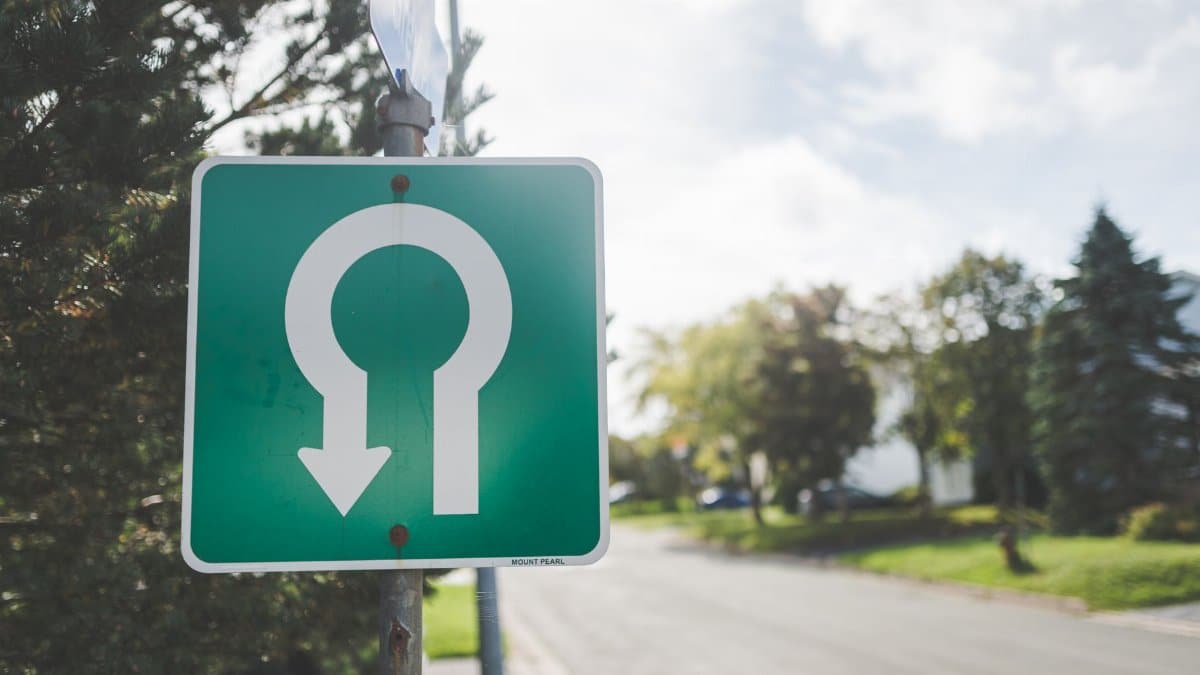 Close-up of a green U-turn road sign on a sunny suburban street with trees.