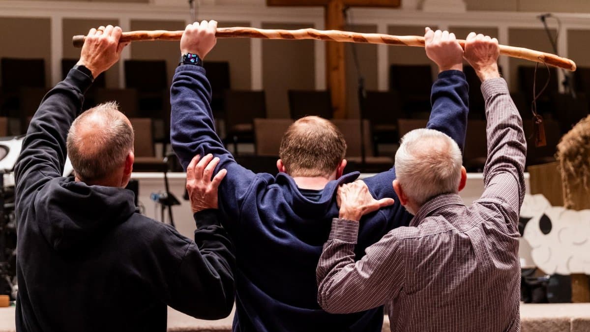 Three men raise a wooden staff in a church setting with a cross visible in the background.