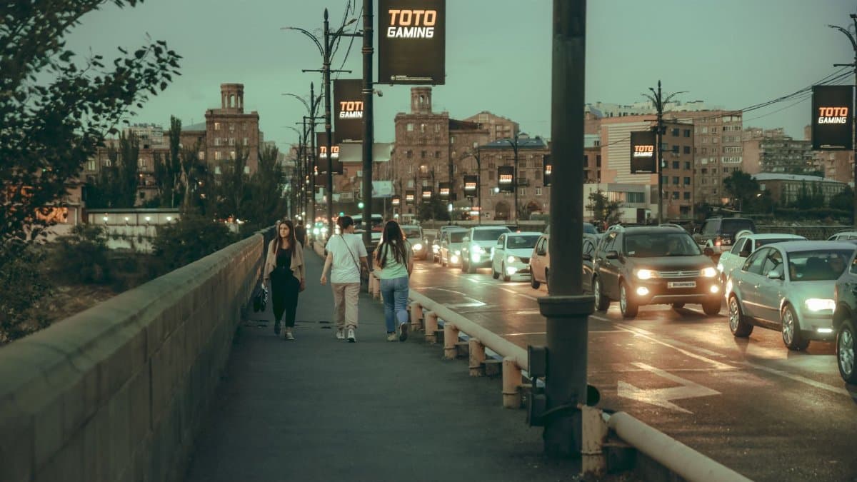 People walk along a busy city sidewalk during evening traffic, with illuminated streetlights.
