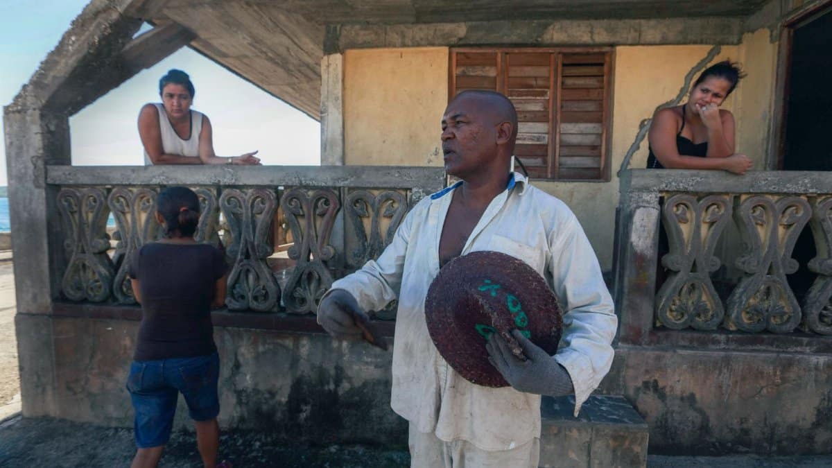 A man holding a woven hat interacts with others outside a rustic house.