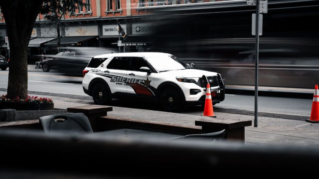 A sheriff vehicle parked on a busy urban street with motion blur of passing cars and vibrant orange cones.