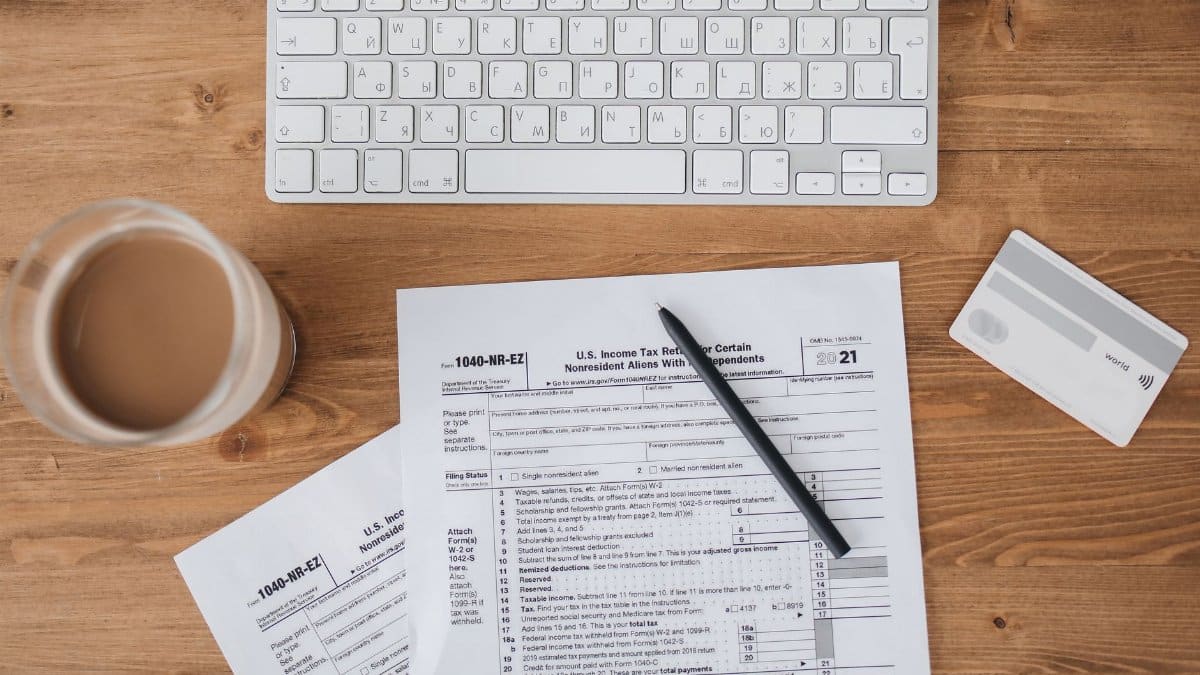 Top view of tax forms, keyboard, pen, and coffee on a wooden desk.