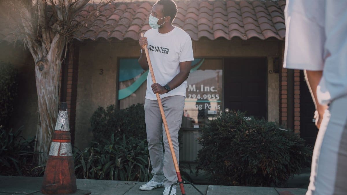 An adult volunteer cleaning a sidewalk with a broom, wearing a face mask for safety.