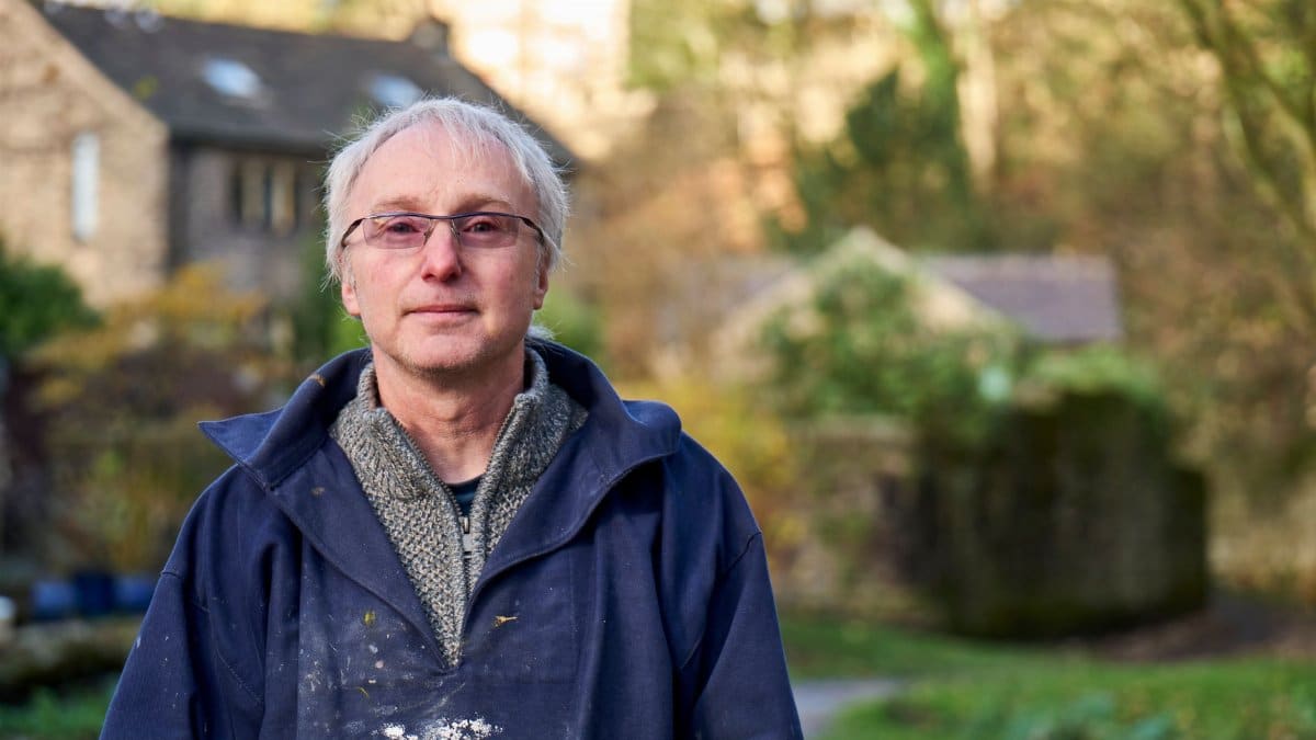 Portrait of a senior man wearing glasses and a blue jacket standing outdoors with a house in the background.