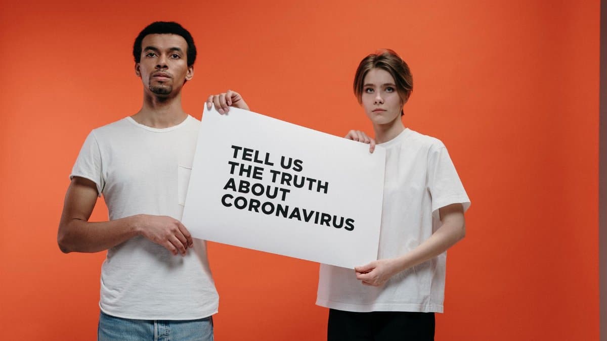 Two people holding a 'Tell Us The Truth About Coronavirus' sign against a vibrant orange background.