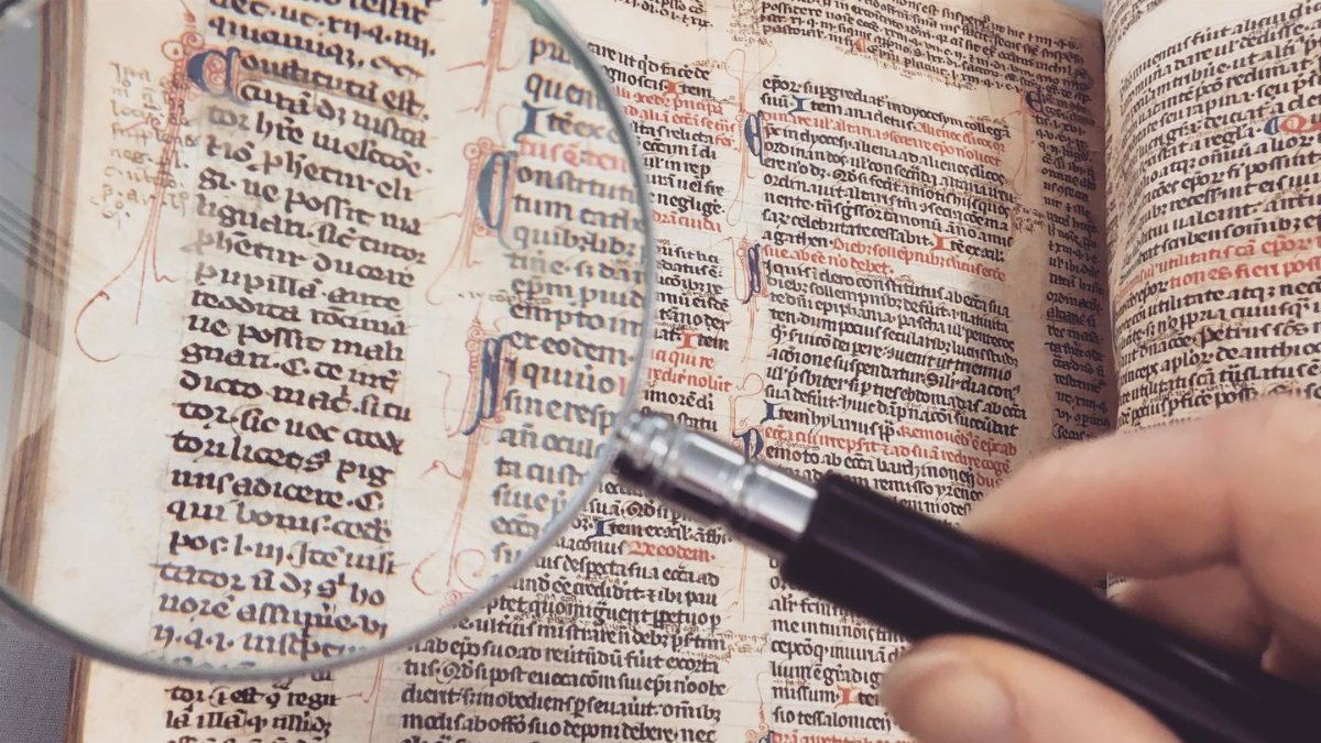 A hand holding a magnifying glass over an antique book page, revealing intricate text and design.