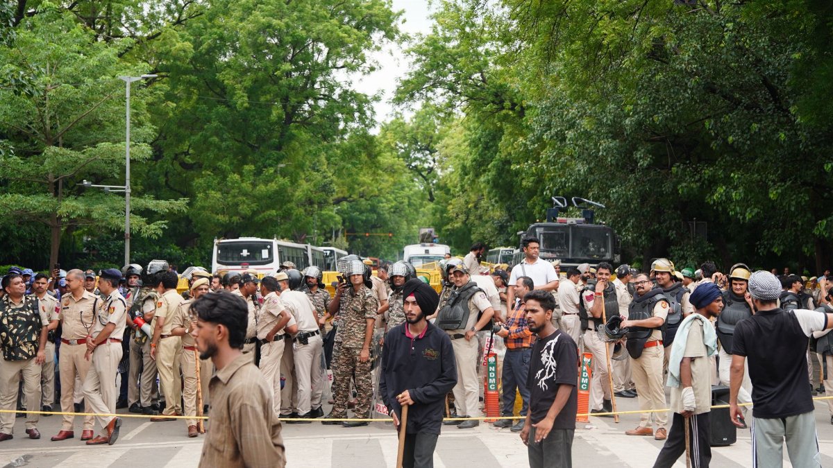 Police line monitors a diverse crowd outdoors amidst greenery and vehicles.