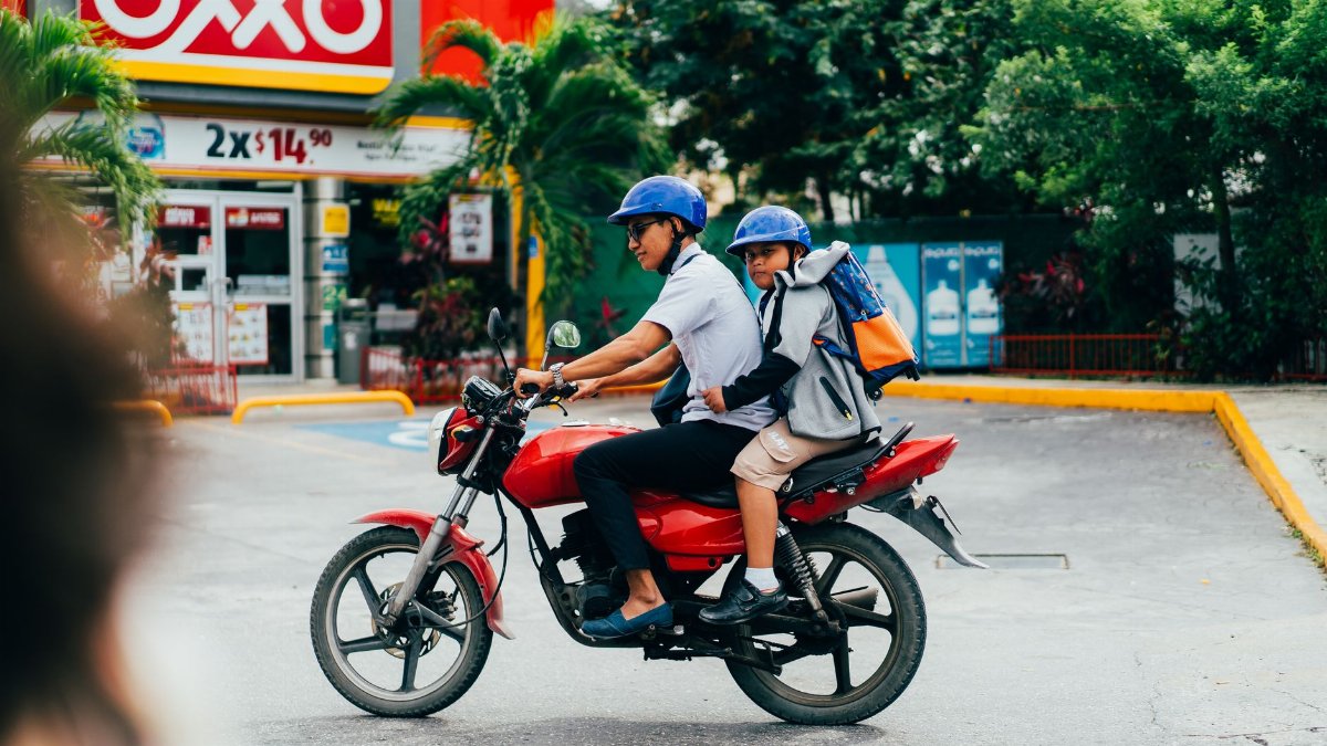 A father and son ride a red motorbike past an OXXO store, showcasing urban transportation.