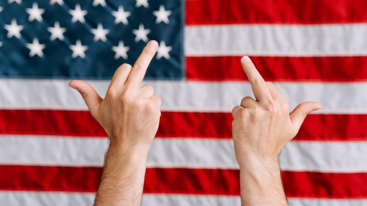 Hands displaying a rebellious gesture in front of the American flag, symbolizing defiance.