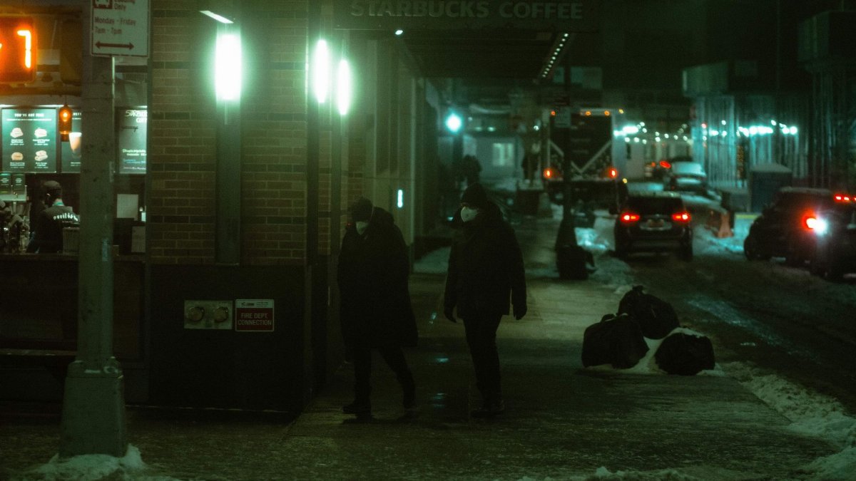 People in protective masks strolling on snowy walkway between residential building and road with cars on dark street at evening time