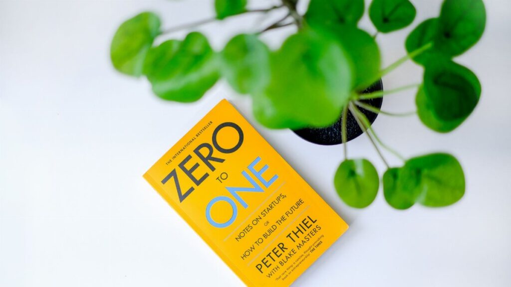 Overhead view of 'Zero to One' book next to a green plant on a white surface.