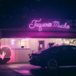 A neon sign illuminates a taqueria with a parked car at night, capturing urban nightlife.