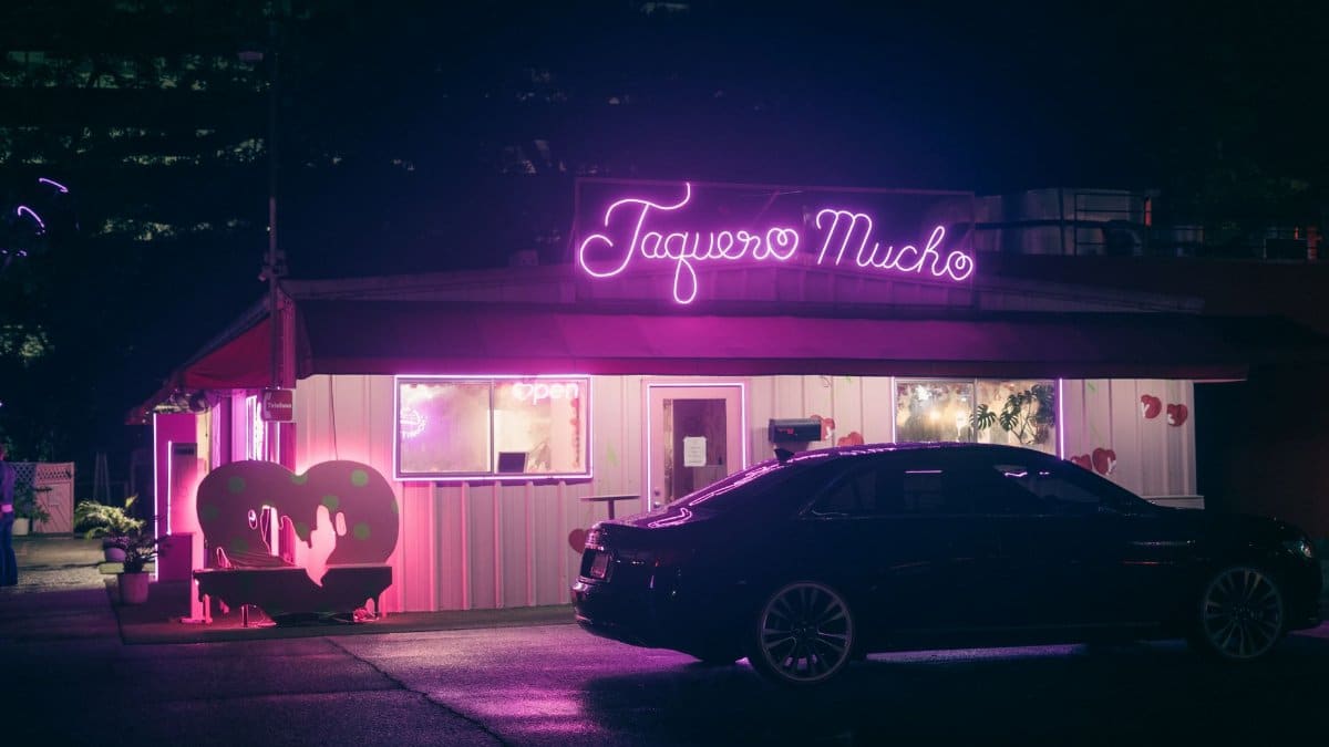 A neon sign illuminates a taqueria with a parked car at night, capturing urban nightlife.