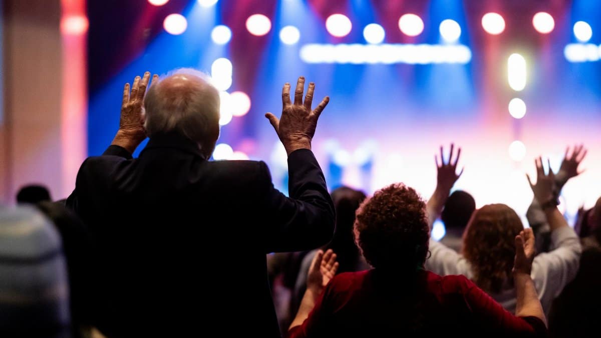 Dynamic scene of a worship concert with people joyfully raising their hands in praise under colorful stage lights.