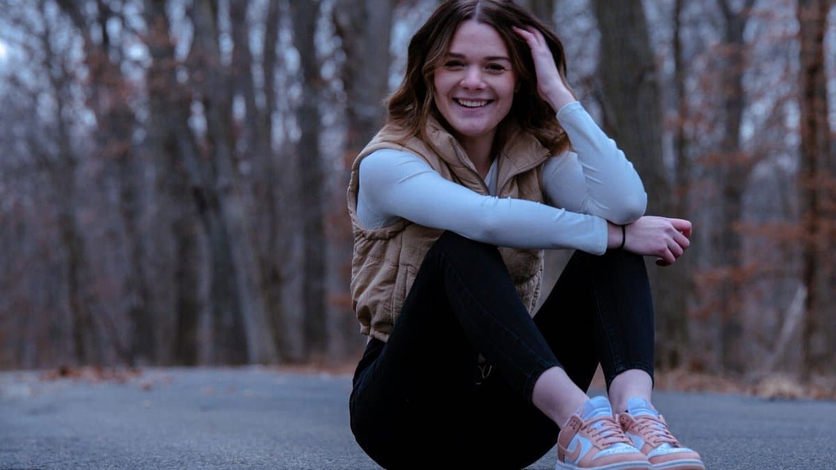Smiling woman in casual attire sits on a forest road in fall setting.