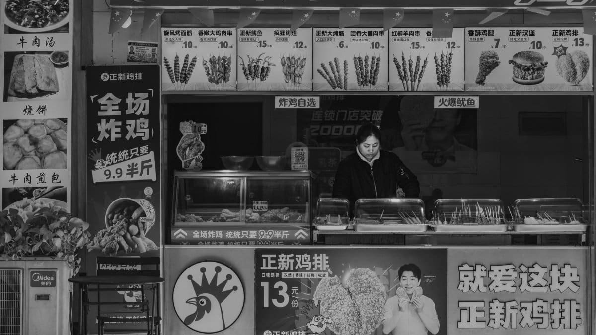 Black and white photo of a street food stall with vendor, displaying various menu items and prices.