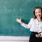 Smiling teacher in a classroom pointing at back to school message on a chalkboard.