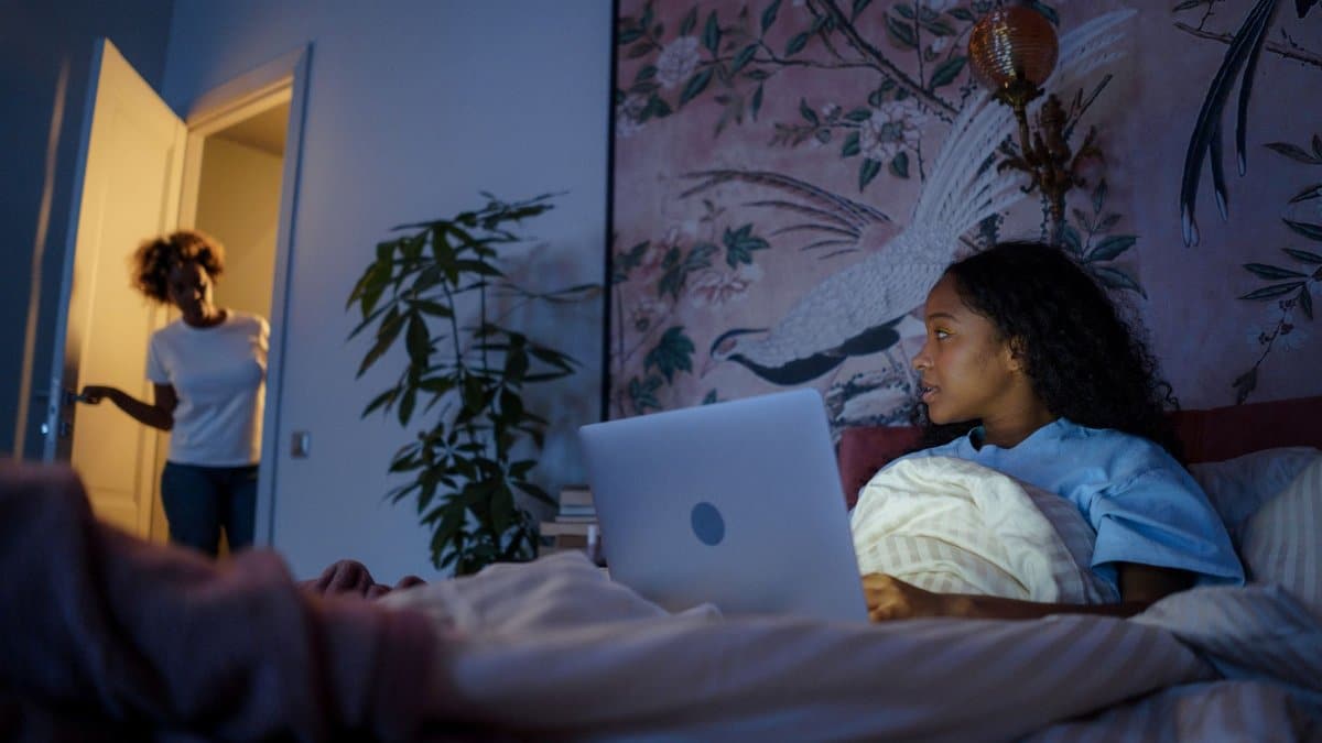 A teenager with a laptop in a dimly lit bedroom looks towards a parent in the doorway.