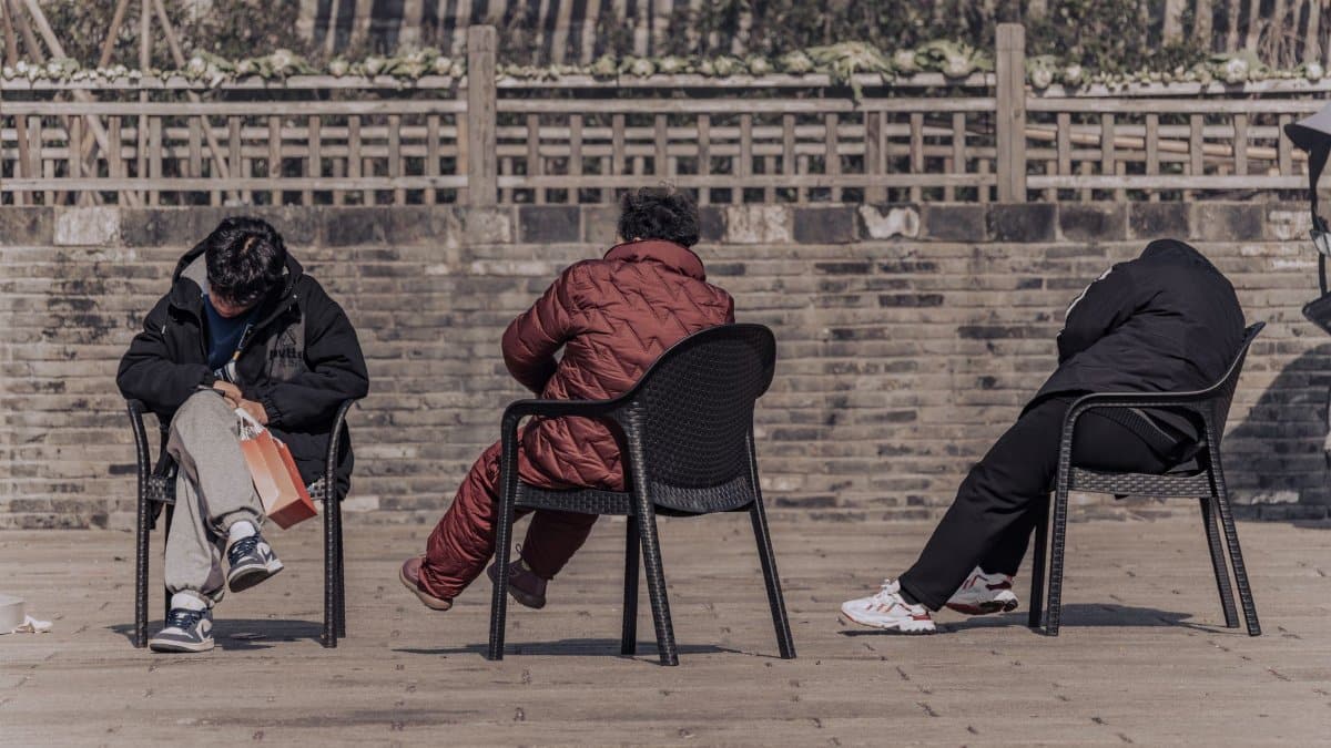 Three adults relax in chairs outdoors against a brick wall, enjoying a sunny day.