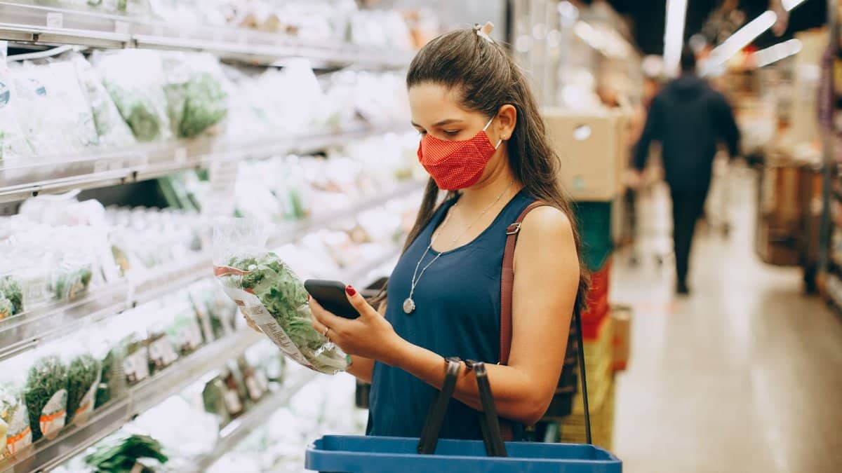 A woman wearing a mask shops for groceries in a supermarket, checking products with her phone.