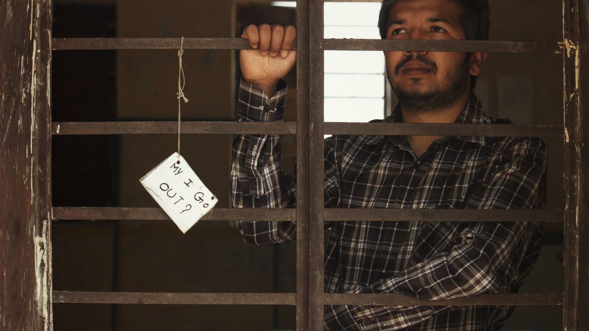 Man standing behind bars holding a sign asking 'May I Go Out?' Indoor setting with dramatic tone.