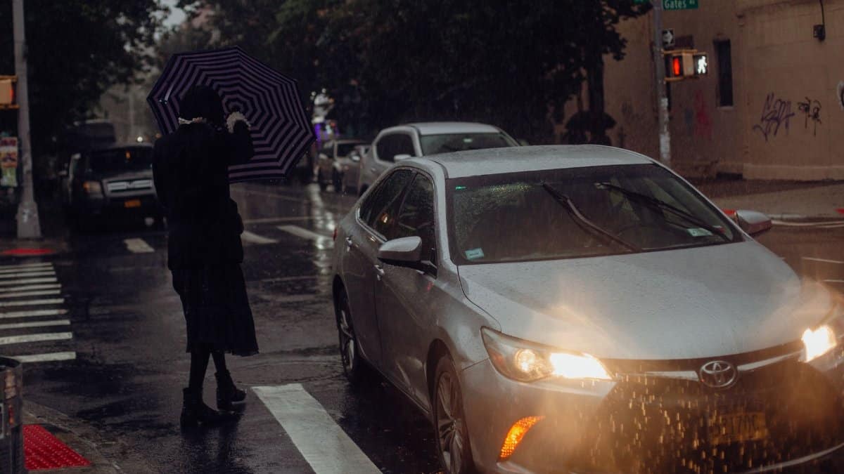 A person with an umbrella crosses a wet urban street in the rain, near a silver car.