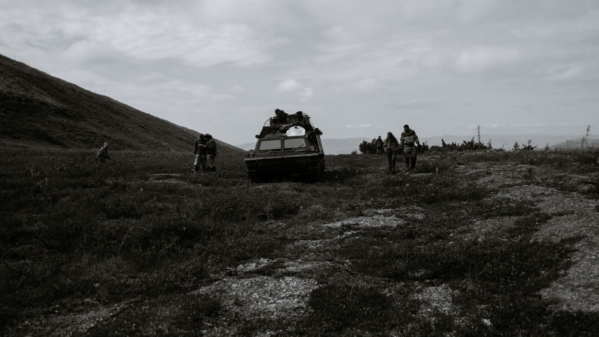 Black and white of anonymous people walking on dry grassy meadow at hillside near tank during military training against overcast sky