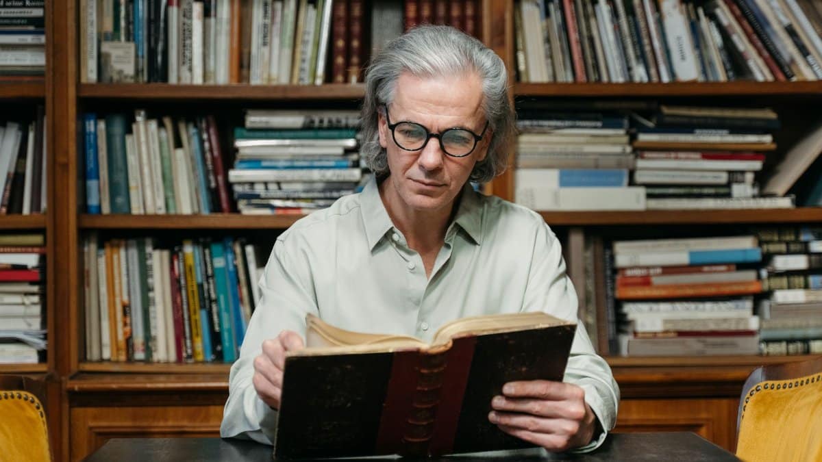 Senior man with eyeglasses reading a book in a library full of bookshelves.