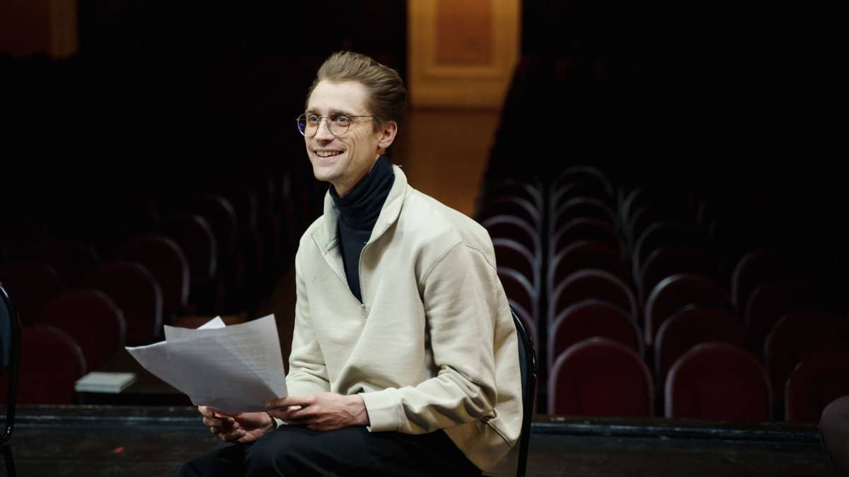 Smiling actor rehearsing script alone in empty theater auditorium, seated on stage.