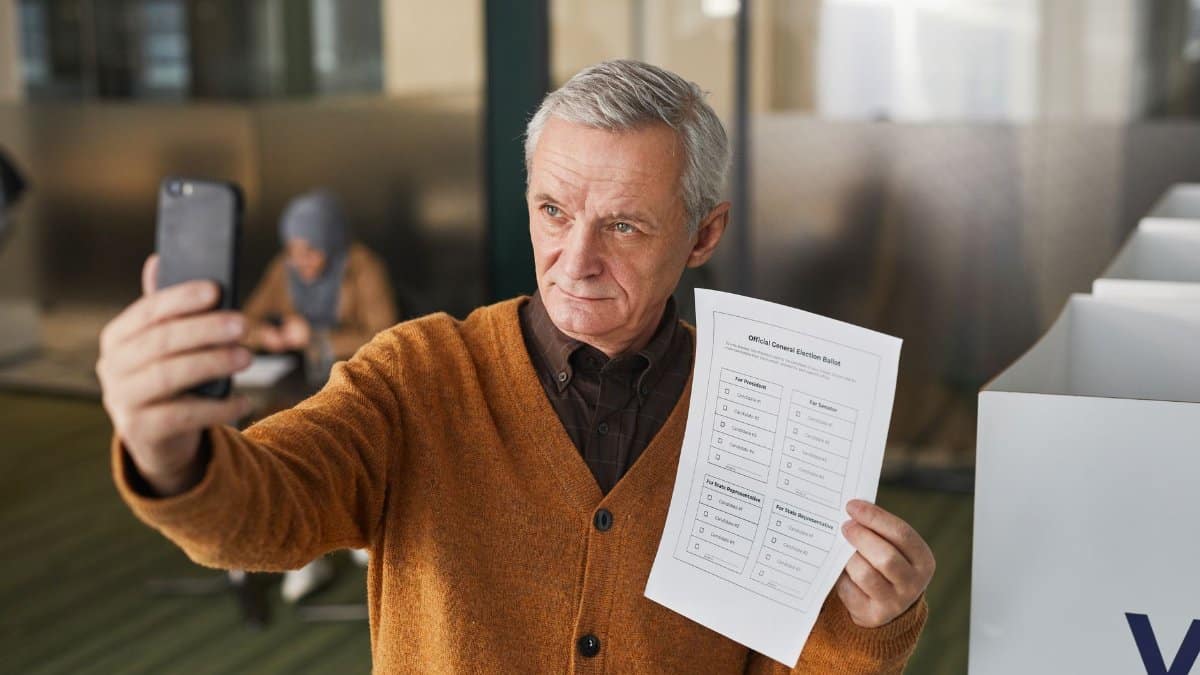 Elderly man with gray hair holding a ballot and taking a selfie in a voting booth.